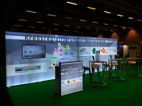 Zu sehen ist der gemeinsame Stand inmitten der Messe. Man sieht einen Standpult und Stühle an Tischen.